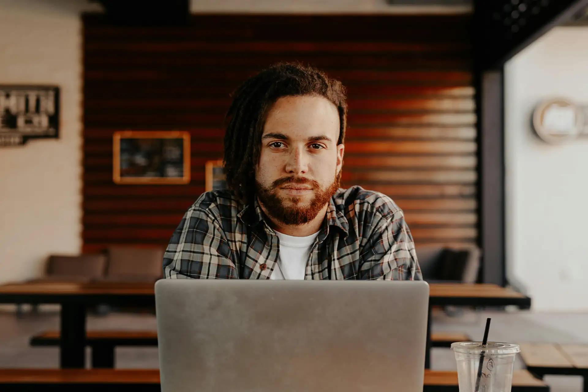 Person mit Bart und kariertem Hemd arbeitet am Laptop in einem gemütlichen Café mit hölzernem Interieur